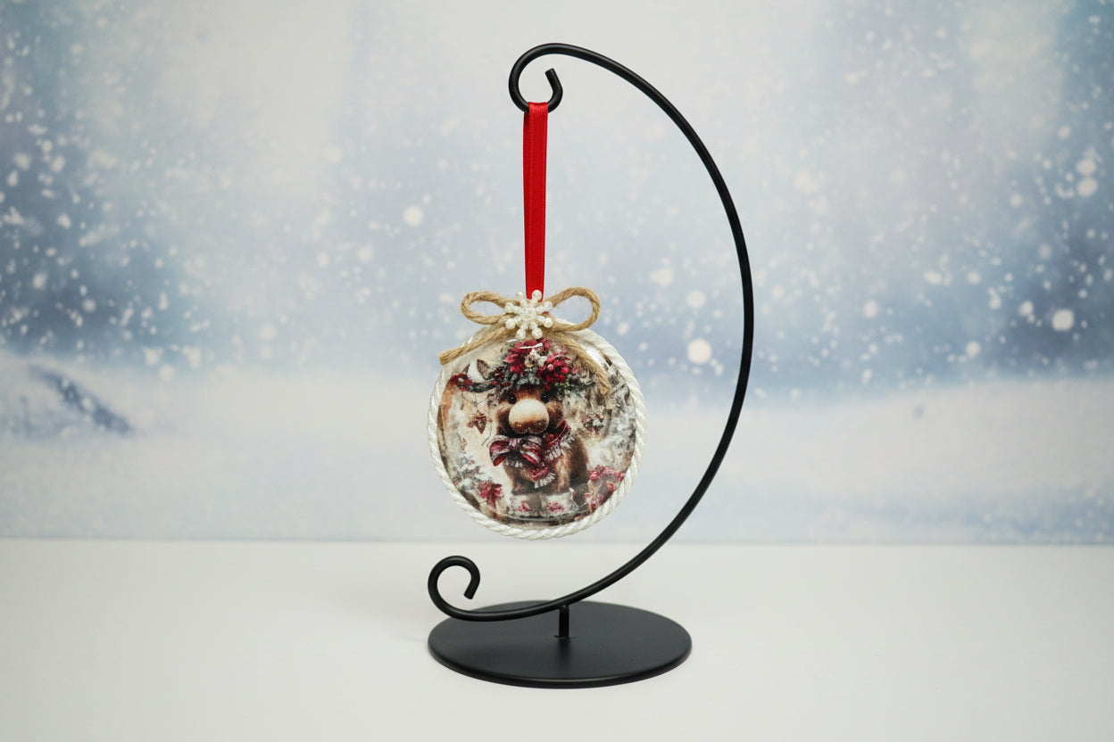 The ornament showcases a cute, illustrated highland-style cow wearing a Santa hat and a festive winter wreath around its neck.