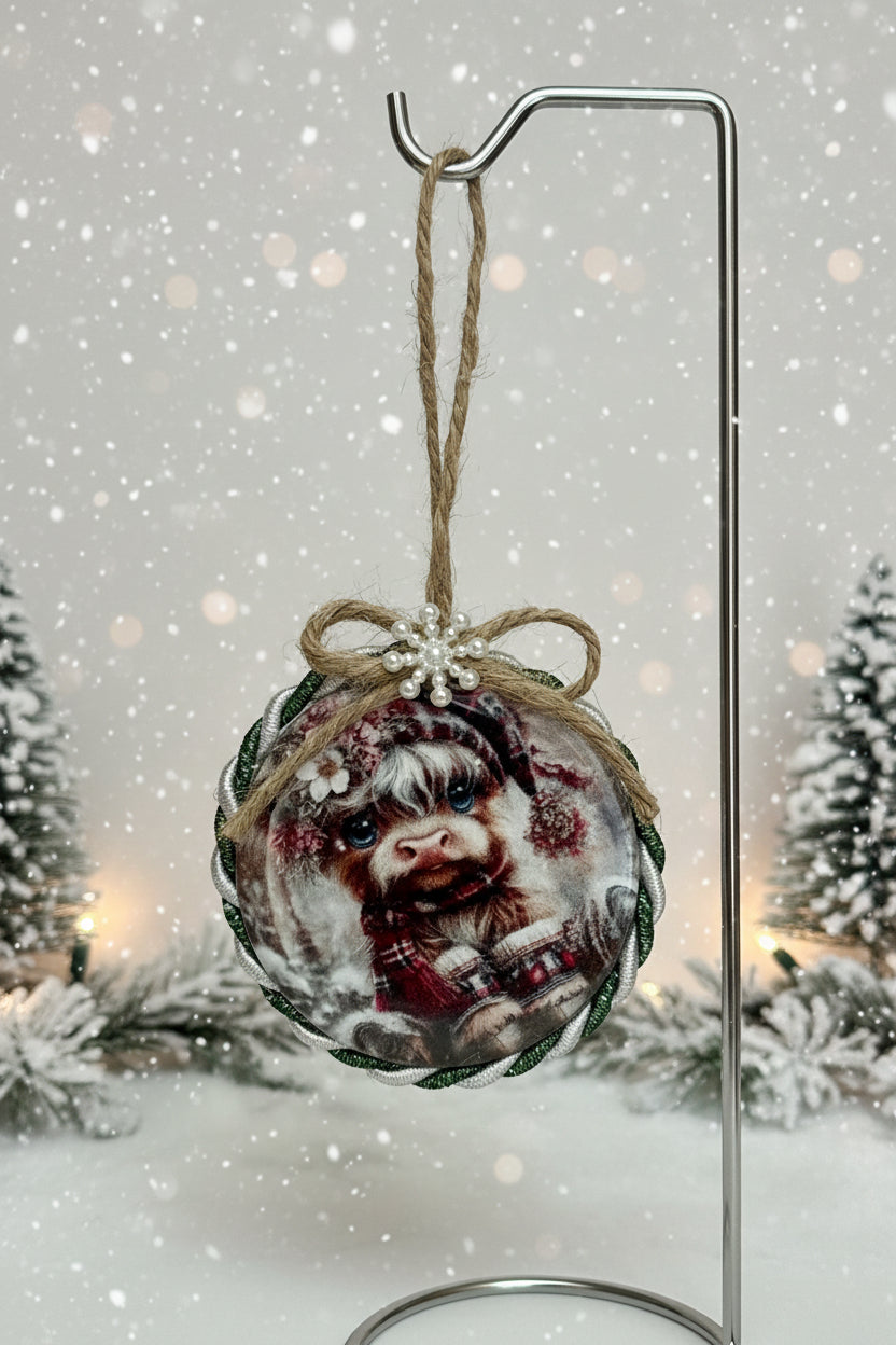 Decorative ornament with a printed design of a Highland Cow wearing a hat and scarf, tied with twine and a pearl-like bead on a plain background.